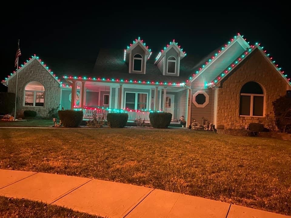 Stone home with alternating red and green Christmas lights on the dormers, roofline and full front porch railing
